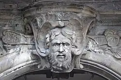 Une sculpture de pierre grise, représentant un visage d'homme barbu, de face.