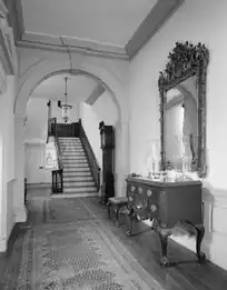 Intérieur d'un hall d'entrée d'une maison historique du XVIIIe ou XIXe siècle, avec des murs blancs, un escalier en arrière-plan, et un sol en bois clair recouvert de tapis géométriques. Un buffet ancien et un miroir classique sont présents. L'image est en noir et blanc.