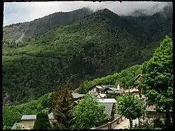 Photographie d'un village qui est enclavé dans sa forêt mais très ensoleillé ; on voit la chapelle au centre du village, entourée de maisons en lauzes et avec la rue arborée.