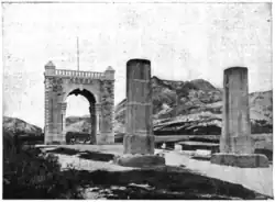 Photographie en noir et blanc montrant à l’avant-plan deux piliers en bois et un arc de triomphe en pierre à l’arrière-plan.
