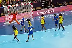 Photo d'une partie de handball où s'affrontent une équipe bleue et une équipe jaune. La gardienne, vêtue de rouge, est en plein saut.