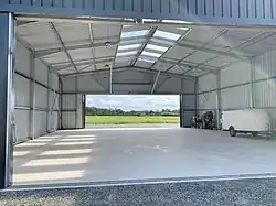 Un des hangars de l’aérodrome : en arrière plan, on aperçoit un biplan Hatz CB-1 en train d’atterrir sur la piste 07, dans le sens sud-ouest vers nord-est.