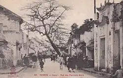 Photographie ancienne montrant des passants dans une rue composée de bâtiments de style extrême-oriental, visiblement mal entretenus.