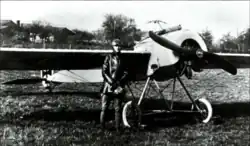 Photo noir et blanc de mauvaise qualité d'un homme en tenue de vol devant un avion monoplan.