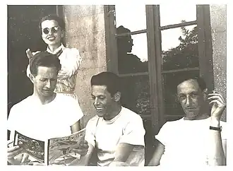Photographie en noir et blanc représentant 3 hommes assis et une femme sourante debout, portant des lunettes de soleil et se livrant à des facéties.