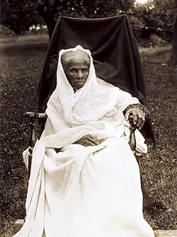 Photographie en noir et blanc d'une femme noire âgée à cheveux blancs, assise dans un fauteuil, tout de blanc vêtue