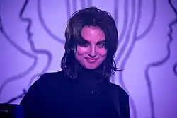 Photo de la chanteuse de Heartworms: une personne souriante, à la peau pâle, brune. L'éclairage est violet et l'arrière-plan présente des lignes intriquées violacées inspirant le profil de deux visages se tenant dos-à-dos.