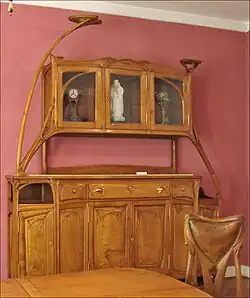 Photo en couleur d'un buffet en bois assez clair et poli avec quelques lignes arrondies, coin de table et dossier de chaise au premier plan