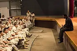 Un homme, assis sur une estrade, micro en main, se tient devant une assemblée de militaires en tenue.