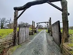 Photo de deux grandes portes en bois à double battant successives, toutes les deux ouvertes. On aperçoit les toits de chaume de maisons rondes à l'arrière-plan