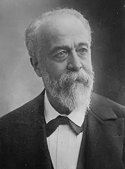 Photo en noir et blanc d’un homme ayant les cheveux blancs, une barbe taillée en pointe et habillé en costume trois pièces, avec un nœud papillon