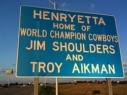 Un panneau bleu avec dessus les inscriptions en lettres blanches suivantes : « Henryetta, Home of World Champion Cowboys Jim Shoulders and Troy Aikman ».