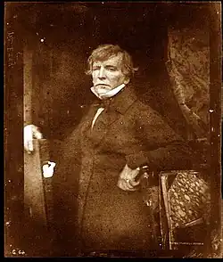 Portrait photographique d'un homme d'âge mûr, de type européen, vêtu à la mode du XIXe siècle. Posture droite, regard légèrement décalé, expression sérieuse. Photographie vintage aux tons brun-beige, éclairage subtil, arrière-plan sombre, composition frontale mettant en valeur le sujet.