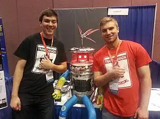 Deux personnes debout encadrent en souriant un robot humanoïde assis sur une table.