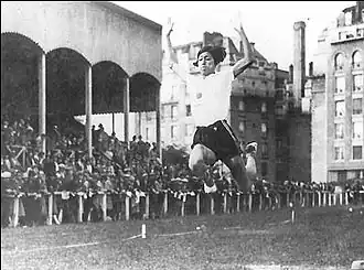 Photo noir et blanc d'une femme en tee-shirt blanc et short noir, saisie dans les airs au cours d'un saut en longueur. Une foule de spectateurs et des bâtiments sont visibles en arrière-plan.