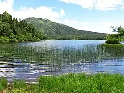 Photo couleur (couleurs dominantes : vert et bleu) d'un étang, avec une montagne en arrière-plan, sous un ciel bleu nuageux.