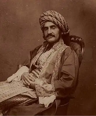 Photographie sépia d'un homme vêtu d'un habit du Moyen-Orient du XIXe&nbsp;siècle, portant une large moustache, assis sur une chaise avec les mains croisées sur ses jambes