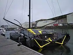 Vue de trois quarts arrière d'un voilier noir et jaune, qui a la particularité d'avoir un cockpit entièrement recouvert et fermé.