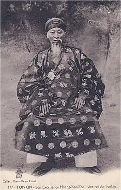 Vieil homme portant un turban, une longue barbe et moustache, une tunique traditionnelle vietnamienne sombre décorée de cercles et de caractères chinois clairs, un pantalon clair et des chaussures sombres. Il est assis.