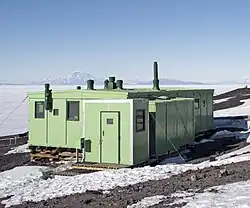 Cabane en tôle verte devant la banquise et en fond un volcan.