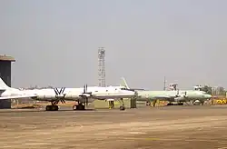 Two four-engine prop-driven aircraft on ramp facing right of screen. The large aircraft in the midground, which is slightly off-centered, has swept-back wings and is painted in a pale grey paints scheme. The other in the background is a straight-wing aircraft painted in pale green