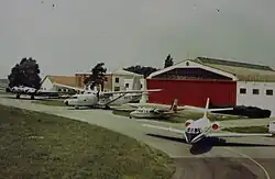 Photographie ancienne représentant quatre avions devant un hangar à la porte rouge. De gauche à droite : un B-17, un Hurel, un AirCommander et un Mystère 20.