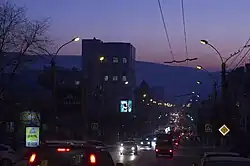 Photographie d'une rue à l'aube avec des voitures et leurs phares. Le trafic est moyennement dense, avec une voie de chaque côté de la rue.