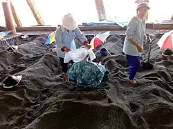 Photo couleur de deux personnes, debout sur un sol sablonneux noire, et d'une troisième personne, de dos, à demi enfouie dans le sable. Des parasols ici et là. Fond lumineux et laiteux.
