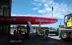 Voilier rouge dont seule la moitié avant est sortie du hangar. Des techniciens ajustent dans son puits un foil blanc qui est soutenu par un chariot élévateur.