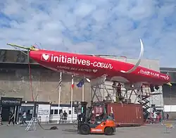 Bateau rouge à foils blancs, sans son mât, posé en hauteur, vu d'en dessous. On voit le haut de la quille, jaune, que des techniciens s'apprêtent à fixer.