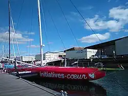 Vue trois quarts avant d'un voilier rouge amarré au ponton. La bôme et les voiles ne sont pas encore en place.