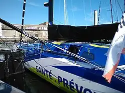 Vue de trois quarts arrière de l'avant d'un voilier bleu, blanc et jaune amarré au ponton.