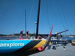 Vu de trois quarts avant, un voilier noir, bleu et jaune amarré au ponton. Bout-dehors et foils rouges. Mât noir.