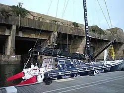 Voilier bleu sombre et blanc, vu de trois quarts arrière, amarré au ponton. En arrière-plan, l'ancienne base de sous-marins allemande de Lorient.