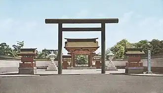 Photo de l'entrée d'un sanctuaire, avec un torii immense à l'entrée, et des lanternes de pierre de chaque côté. Au fond de l'image, on devine le bâtiment principal au bout d'une longue allée rectiligne.