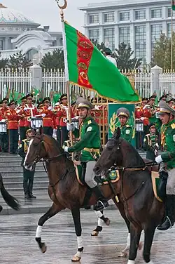 Cavalier sur un cheval en marche tenant un drapeau, suivi par deux autres soldats à cheval. En arrière-plan, une fanfare se tient devant les grilles d'un grand bâtiment blanc.