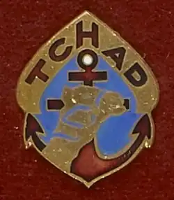 Un insigne présentant : une ancre de gueules (rouge) chargée d'un chameau d'or, en chef l'inscription Tchad dans une arche d'or.