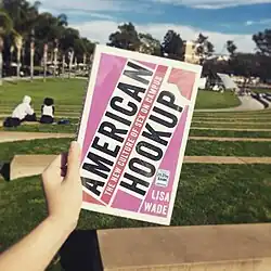 Une main tient un livre fermé à couverture rose. On voit derrière le livre un parc ensoleillé.