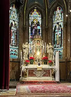 Photographie couleur montrant l'autel décoré de sculptures et de peintures et derrière des grands vitraux lumineux représentants des personnages en pied.