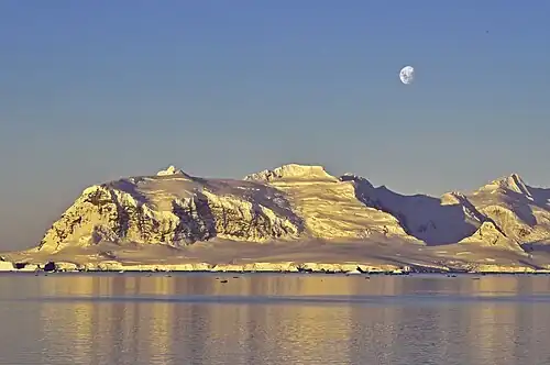 Photographie en couleurs d'un paysage montagneux faiblement éclairé, avec la lune visible en haut et à droite.