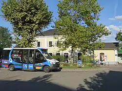 Photographie en couleurs d’un minibus bleu, blanc et noir stationné devant la gare ferroviaire de Rumilly, en arrière-plan.
