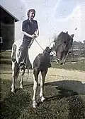 Photographie colorisée. Au centre, Janet Rosenberg coiffée et vêtue à la garçonne montant un cheval à la robe pie tovero. Le cheval est à l'arrêt. Le paysage est rural et le ciel dégagé.