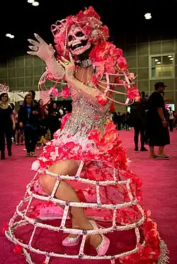 Drag queen sur un tapis rouge. Son visage est recouvert d'un masque de squelette entouré de fleurs roses et de perles. Elle porte des épaulières composées uniquement de leur structure et recouvertes de roses, un corset perlé, des gants rose pale de satin et une jupe rigide dont on ne voit que la structure recouverte de perles. Au second plan, plusieurs personnes en tenues sombres se baladent.