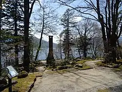 Photo couleur d'un jardin boisé au bord d'une étendue d'eau, sous un ciel bleu nuageux.
