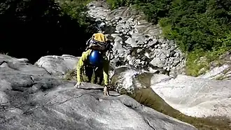 Photo couleur d'un homme grimpant paroi rocheuse grise le long de laquelle s'écoule en chute l'eau d'un ruisseau