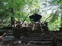 Photo couleur d'un édifice religieux en bois noir, au sommet d'un escalier en pierre et entouré d'arbres sous un ciel nuageux.