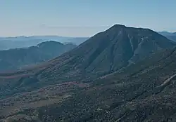 Photo couleur montrant une montagne aux pentes boisées et un massif montagneux au second plan, sous un ciel bleu.