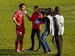 Une femme interview un joueur de rugby, entourée par son personnel technique.