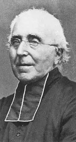 Photographie en noir et blanc d'un homme habillé en soutane noire avec des lunettes et des cheveux blancs.