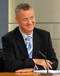 Un homme portant des lunettes et en costume, assis, souriant, devant un micro posé.